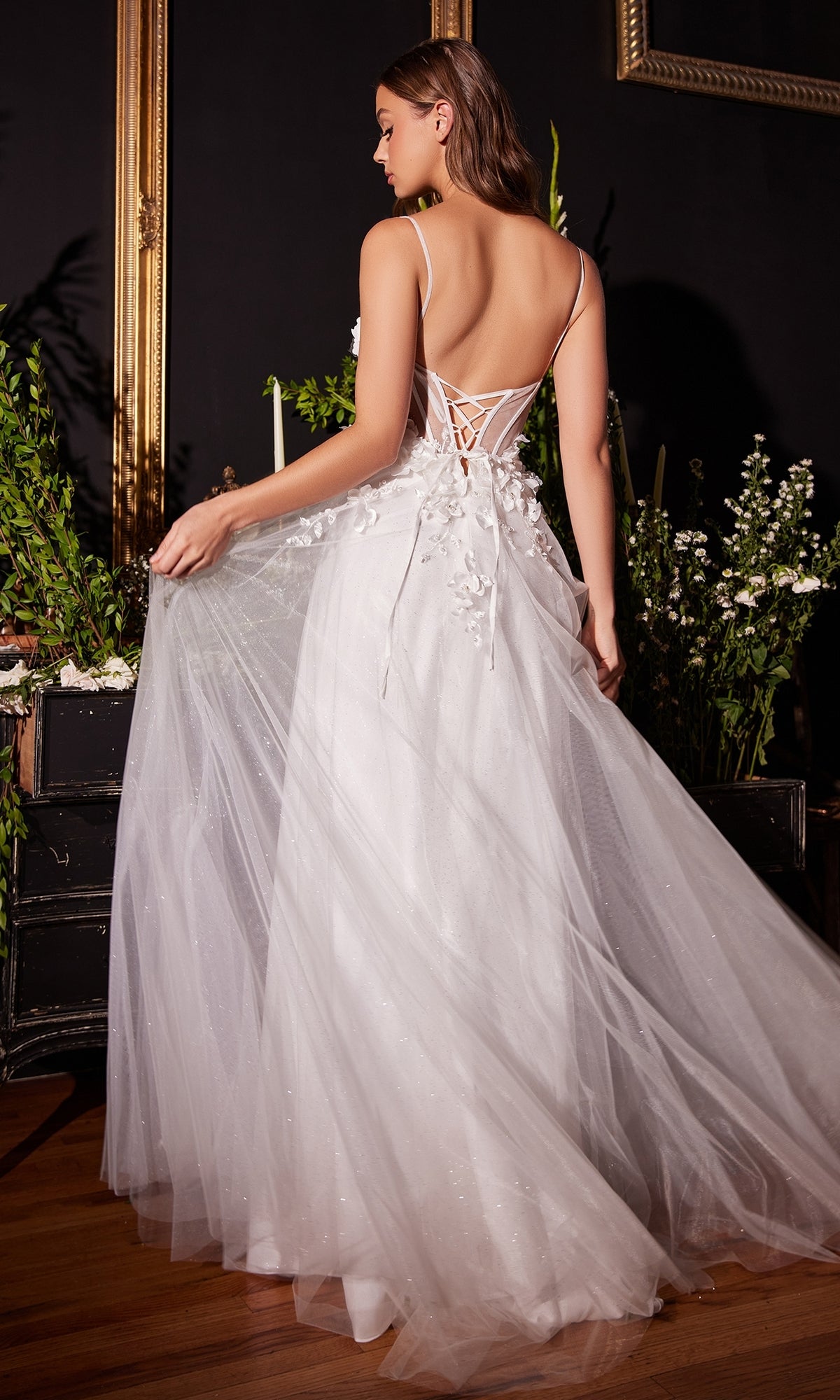A woman indoors holds out the floral-embellished skirt of her Off-White Long Floral Prom Ball Gown CM321W, featuring a lace-up back and 3-D flowers, surrounded by greenery.